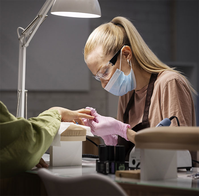 Woman providing nail service, wearing gloves and mask, focused on the task under bright lamp.