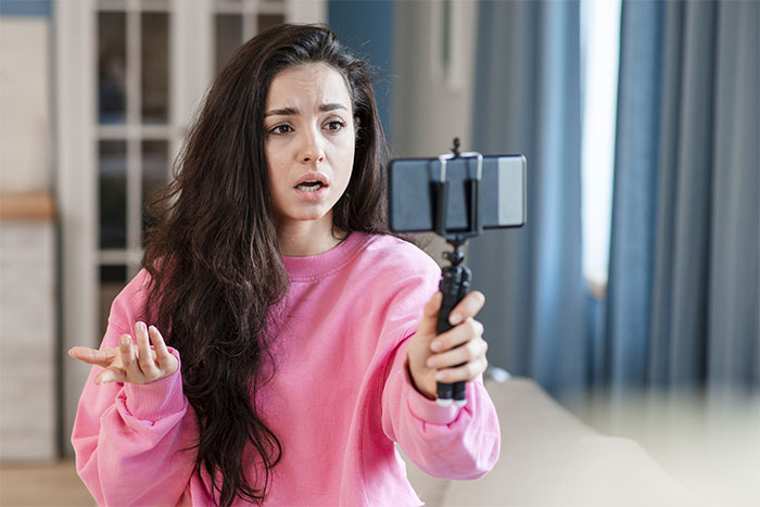 Woman in a pink sweater filming herself with a smartphone, discussing trading services.
