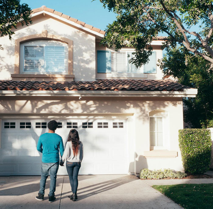 A couple stands in front of a suburban house under a clear sky, suggesting financial stability and family dynamics.