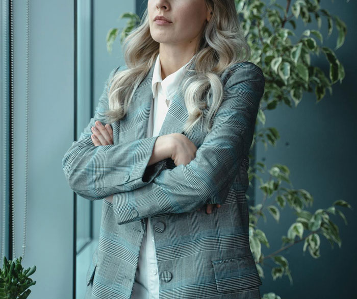 A woman in a plaid blazer with crossed arms, standing by a window, looking thoughtful.