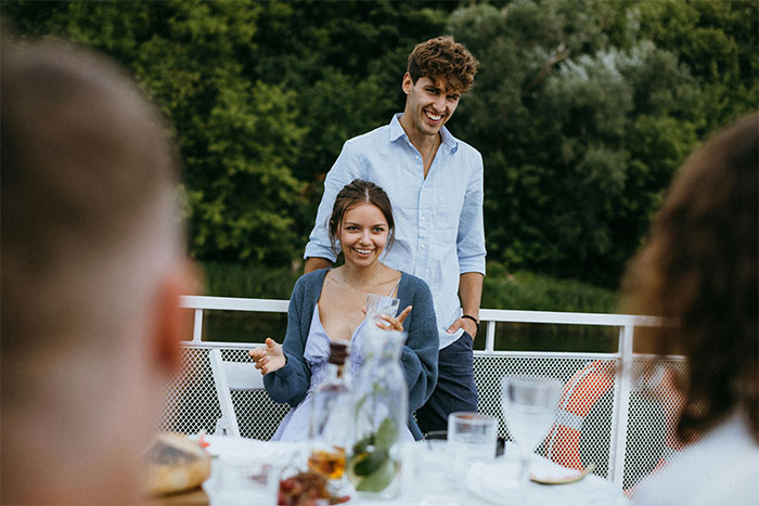 A couple smiling at an outdoor dinner, surrounded by friends, amidst a discussion about paying for everyone's meal.