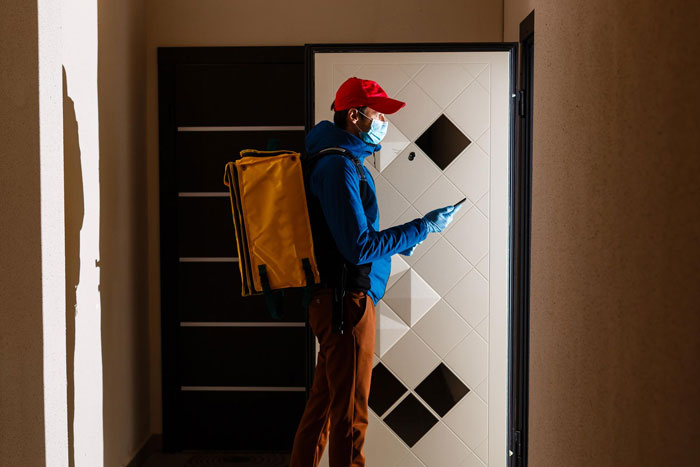 Delivery company driver with a yellow bag at a white door, wearing a red cap and mask, in a dimly lit hallway.