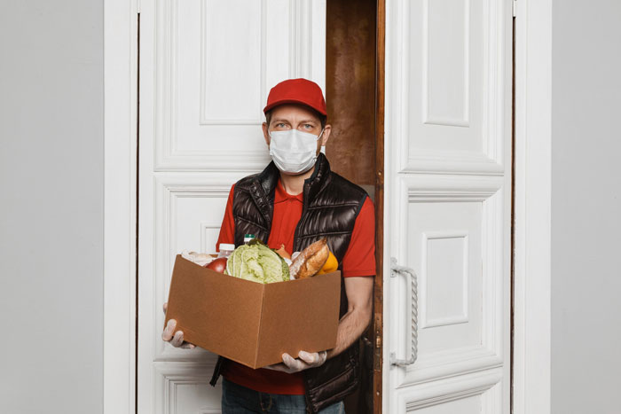Delivery driver in a red cap and mask holding a grocery box at an open front door.