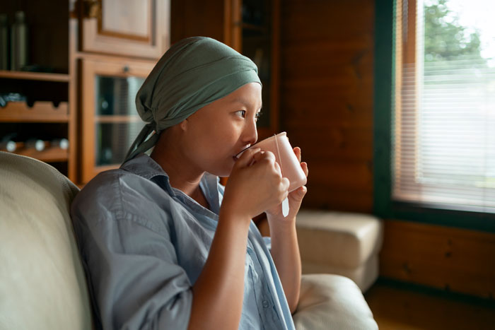 Employee with cancer diagnosis, wearing headscarf, sips from a mug on a cozy couch by a window.