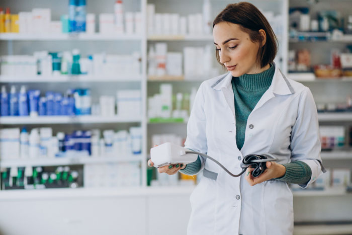 Pharmacy manager in a white coat examining a medical device, surrounded by shelves of medication.