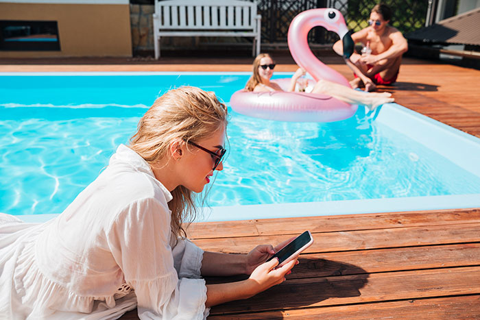 Woman in sunglasses using phone by pool while neighbors’ relatives enjoy pool with inflatable flamingo nearby.