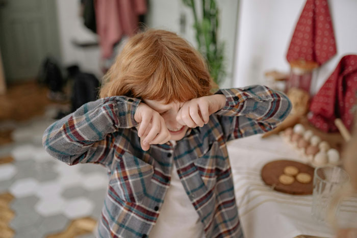 Young red-haired child in plaid shirt wiping eyes indoors, illustrating impact of wealth flexing on childhood experiences.