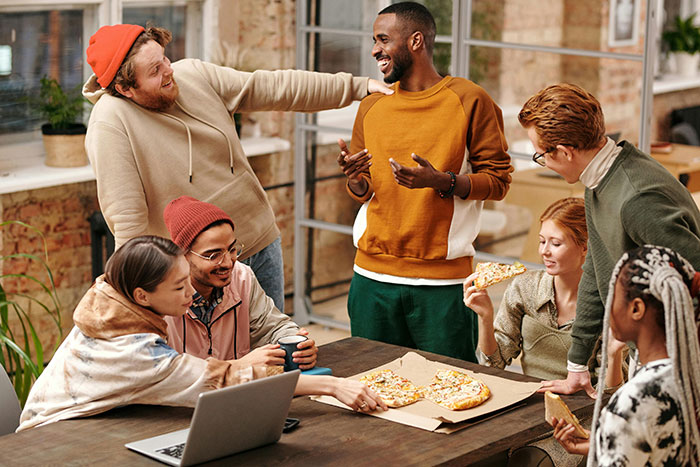 Group enjoying pizza and laughter, person in white hoodie making jokes, lively discussion scene.