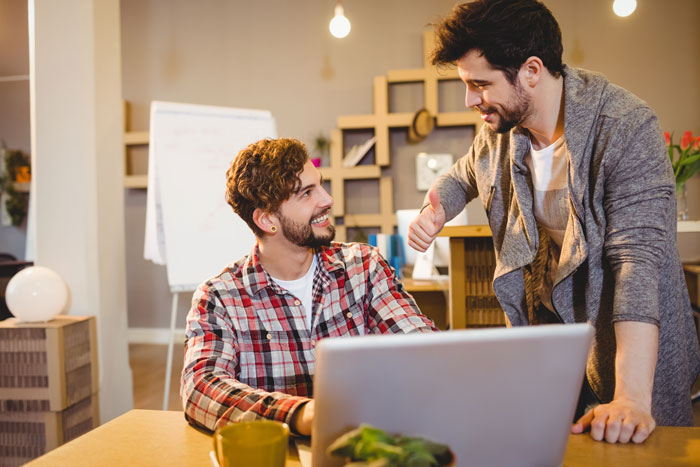 Two coworkers in a discussion at the office, one seated with a laptop, exchanging emails about a recent events at work.