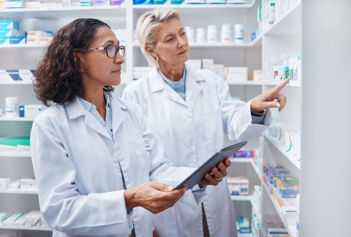 Two female coworkers in lab coats discussing medication on pharmacy shelves, highlighting rude woman and coworker tension.