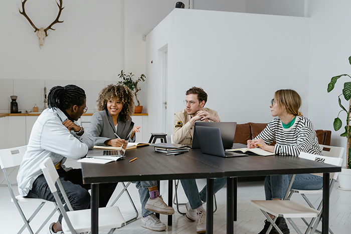 A group of employees discussing work around a table in a modern office, highlighting lazy employee and new boss dynamics.