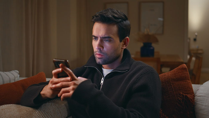 Man sitting indoors, looking at a smartphone with a focused expression.