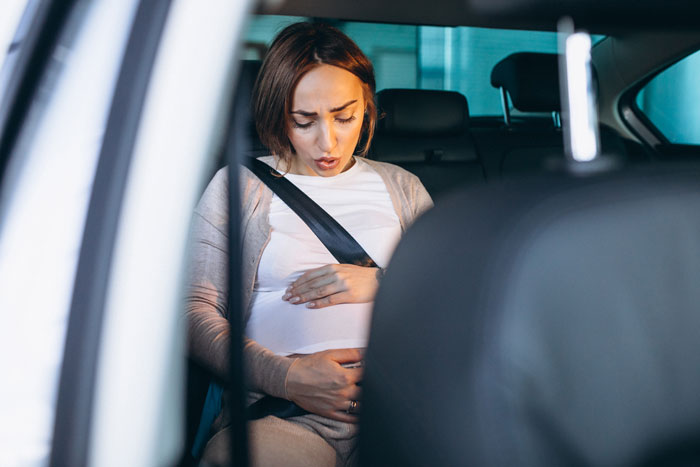 Pregnant neighbor in car, clutching belly, prepared for ride with waterproofed seats for protection.