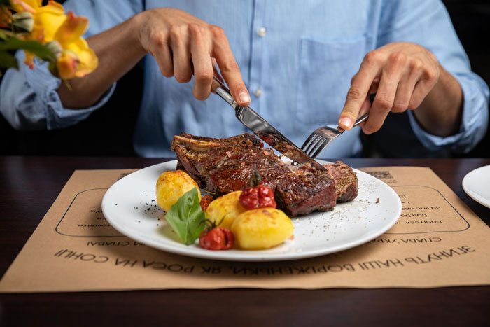Person cutting steak with fork and knife, highlighting friend laughs off vegan’s warning about paying for his food.