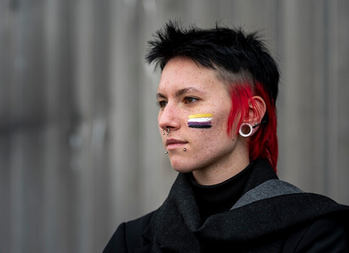 Person with short black and red hair and nonbinary pride flag face paint, representing themes of trans identity and self-discovery.